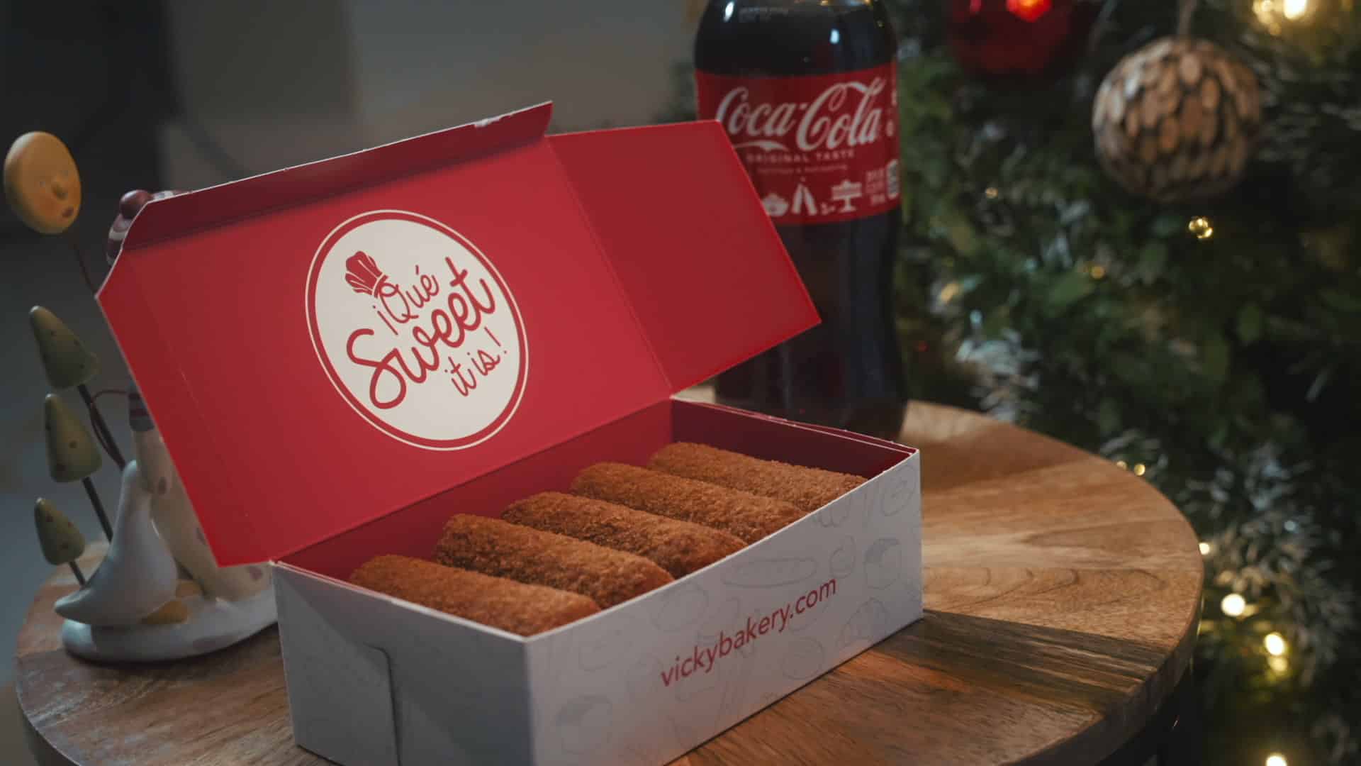 Open red and white box from Vicky Bakery filled with croquetas, placed on a wooden table next to a Coca-Cola bottle and Christmas decorations.